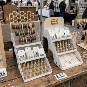 Könnte beinhalten: Zwei weiße, mehrstufige Schmuckständer mit verschiedenen Schmuckstücken. Die Ständer halten Armbänder, Ohrringe, Ringe und Halsketten. Die Ständer haben eine Holzplatte mit dem Text "EK DESIGN". Der Schmuck wird auf einem Holztisch ausgestellt.