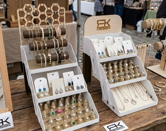 May include: Two white tiered jewelry display stands with various jewelry items. The stands hold bracelets, earrings, rings, and necklaces. The stands have a wooden top with the text "EK DESIGN". The jewelry is displayed on a wooden table.