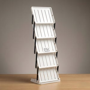 May include: A white, multi-tiered display stand with black hinges. Each tray has vertical slots. The stand has the text "RK DESIGN" on one of the trays. The stand is on a wooden surface.