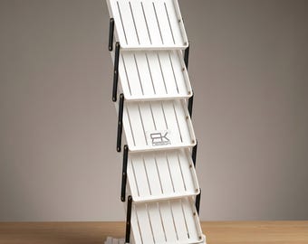 Könnte beinhalten: Ein weißer, zusammenklappbarer Prospektständer mit fünf gestuften Ablagen, schwarzen Metallseitenstützen und einem Logo mit der Aufschrift RK DESIGN auf der mittleren Ablage. Das Gerät ist vertikal ausgerichtet und hat geschlitzte Oberflächen.
