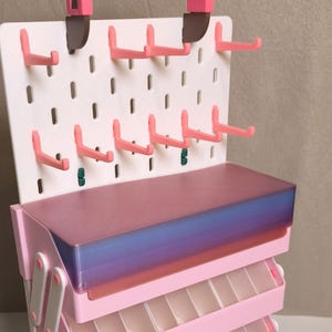 May include: A pink and white tiered storage organizer featuring a pegboard back panel with multiple pink hooks and a top handle. The unit includes several stacked, pull-out trays with translucent, gradient-colored lids that transition from purple to blue.