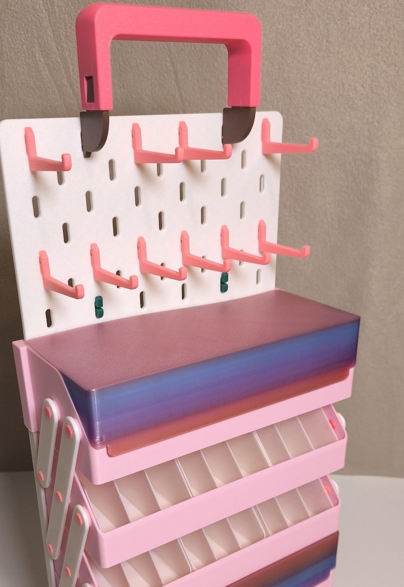 May include: A pink and white tiered storage organizer featuring a pegboard back panel with multiple pink hooks and a top handle. The unit includes several stacked, pull-out trays with translucent, gradient-colored lids that transition from purple to blue.