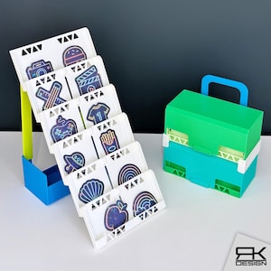 May include: A white tiered display stand with blue and lime green supports, holding several square cards featuring neon-style graphics of a camera, burger, controller, jersey, fries, UFO, seashell, apple, and rainbow. Beside it sits a green and teal storage box.