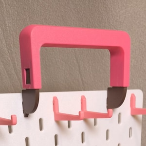 May include: A white pegboard panel featuring multiple pink L-shaped hooks and a large pink carrying handle attached to the top with dark brown brackets. The pegboard has a grid of vertical slots for organizing items, with the handle centered.