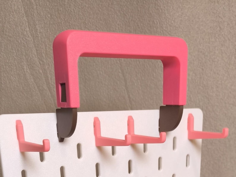May include: A white pegboard panel featuring multiple pink L-shaped hooks and a large pink carrying handle attached to the top with dark brown brackets. The pegboard has a grid of vertical slots for organizing items, with the handle centered.