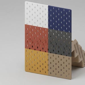 May include: A rectangular pegboard panel featuring a grid of vertical slots, divided into six distinct color sections: white, dark blue, terracotta, dark gray, mustard yellow, and beige, supported by a beige plastic stand on the right side.