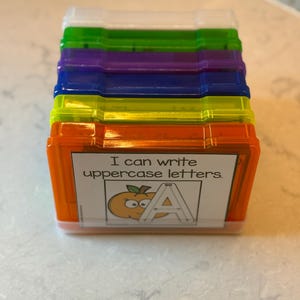 May include: A stack of colorful plastic boxes, each with a clear lid. The boxes are orange, yellow, blue, purple, green, and white. The front of the orange box has a label that reads "I can write uppercase letters."