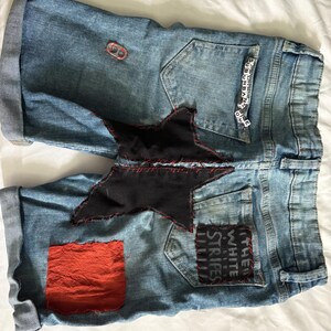 May include: Light blue denim shorts with rolled cuffs. Features a large black star patch with red stitching, a red square patch, and a patch that says "THE WHITE STRIPES". Includes a small metal ring and beads that spell "SUBURBAN HOME".