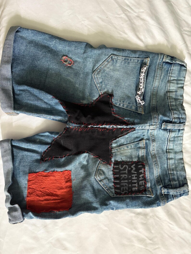 May include: Light blue denim shorts with rolled cuffs. Features a large black star patch with red stitching, a red square patch, and a patch that says "THE WHITE STRIPES". Includes a small metal ring and beads that spell "SUBURBAN HOME".