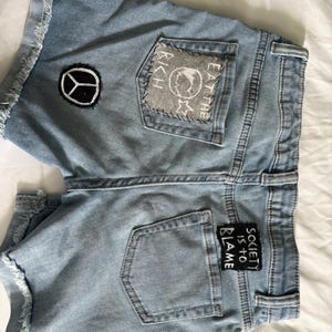 K&ouml;nnte beinhalten: Hellblaue Jeansshorts mit ausgefransten Kanten und Aufn&auml;hern. Ein Aufn&auml;her zeigt ein Friedenszeichen, ein anderer lautet "EAT THE RICH" mit einem Globus und Sternen, und ein dritter sagt "SOCIETY IS TO BLAME".