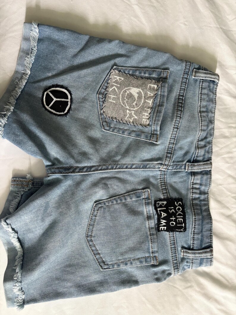 K&ouml;nnte beinhalten: Hellblaue Jeansshorts mit ausgefransten Kanten und Aufn&auml;hern. Ein Aufn&auml;her zeigt ein Friedenszeichen, ein anderer lautet "EAT THE RICH" mit einem Globus und Sternen, und ein dritter sagt "SOCIETY IS TO BLAME".