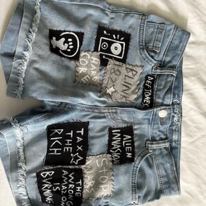 K&ouml;nnte beinhalten: Hellblaue Jeansshorts mit ausgefransten Kanten und verschiedenen Aufn&auml;hern. Aufn&auml;her zeigen Text und Grafiken, darunter "Alien Invasion", "Tax the Rich" und "The Wrong Amazon is Burning". Die Shorts haben einen Knopfverschluss und Taschen.