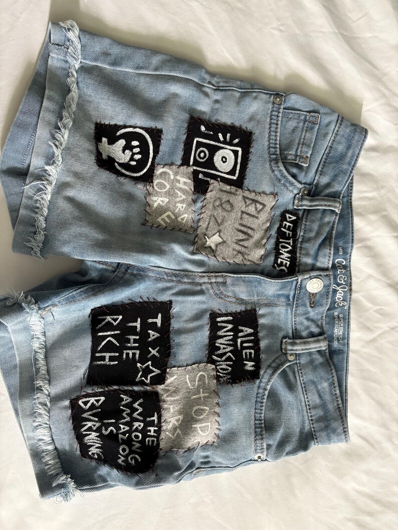 K&ouml;nnte beinhalten: Hellblaue Jeansshorts mit ausgefransten Kanten und verschiedenen Aufn&auml;hern. Aufn&auml;her zeigen Text und Grafiken, darunter "Alien Invasion", "Tax the Rich" und "The Wrong Amazon is Burning". Die Shorts haben einen Knopfverschluss und Taschen.