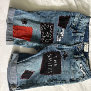 May include: Light blue denim shorts with rolled cuffs. Patches of red and black fabric are sewn onto the shorts, with text and star designs. The text includes "THE SMITHS" and "BEAUTY IS NOT A SIZE".