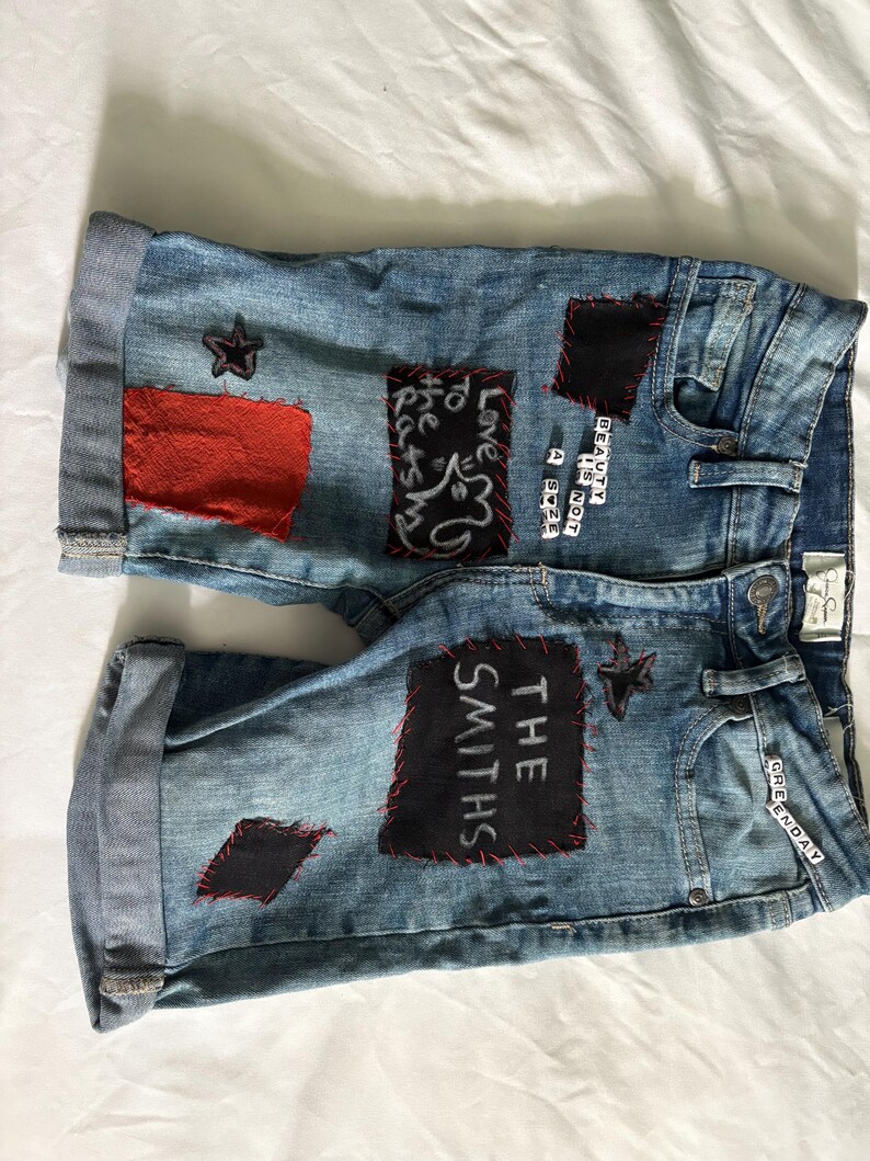 May include: Light blue denim shorts with rolled cuffs. Patches of red and black fabric are sewn onto the shorts, with text and star designs. The text includes "THE SMITHS" and "BEAUTY IS NOT A SIZE".