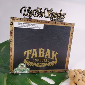Puede incluir: Una caja de madera negra para cigarros con ribetes dorados y una etiqueta dorada que dice "TABAK ESPECIAL". La caja tiene una etiqueta verde con el texto "GUARANTEE UNA CALIDAD INCOMPARABLE".
