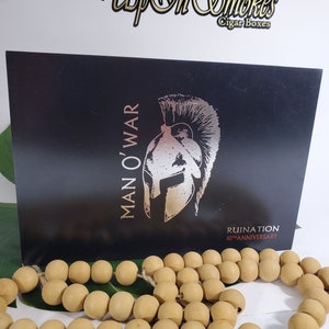 Op de afbeelding: Een zwarte doos met een zilveren Spartaans helm-afbeelding en de tekst "Man O' War" en "Ruination 10th Anniversary". De doos staat op een wit oppervlak met een touwtje van houten kralen.