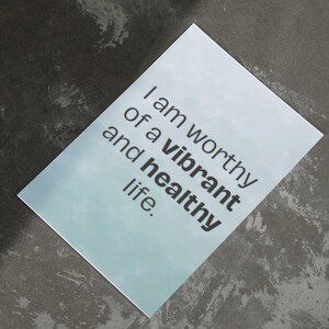 May include: A rectangular card with the text "I am worthy of a vibrant and healthy life" in black font. The card has a light blue gradient background and is set against a dark gray, textured surface.