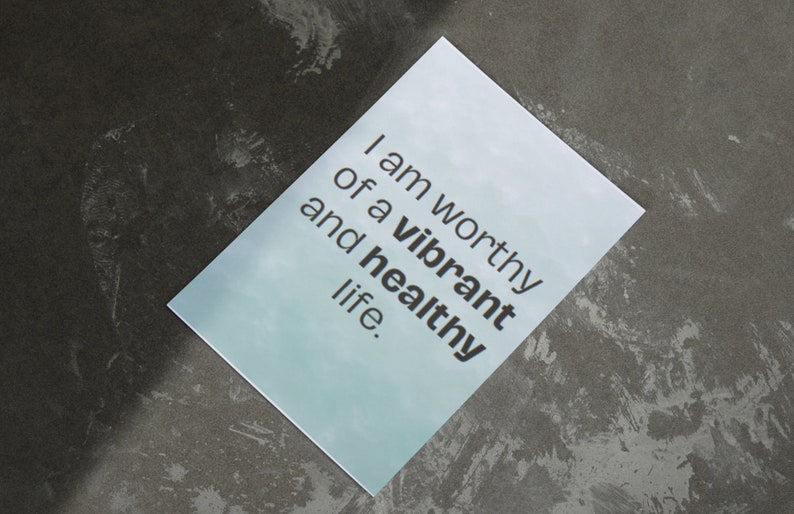 May include: A rectangular card with the text "I am worthy of a vibrant and healthy life" in black font. The card has a light blue gradient background and is set against a dark gray, textured surface.