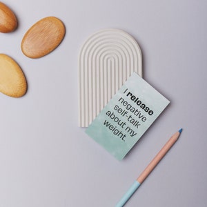 May include: A light gray, arched decorative piece with a series of parallel lines, a teal card with the text "I release negative self-talk about my weight," and a two-toned pencil. Also, three wooden oval-shaped objects.