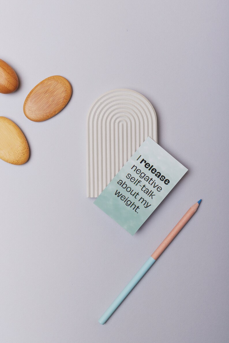 May include: A light gray, arched decorative piece with a series of parallel lines, a teal card with the text "I release negative self-talk about my weight," and a two-toned pencil. Also, three wooden oval-shaped objects.