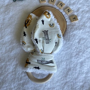 Puede incluir: Un sonajero de conejo de tela blanca con un anillo de madera. La tela tiene un estampado de bosque con árboles, arcoíris y flechas. La palabra "BUNNY" está escrita con fichas de letras.