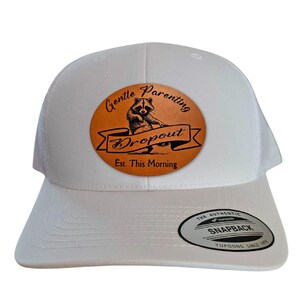 May include: White trucker hat with a curved brim and a mesh back. The front features a circular patch with a raccoon illustration and the words "Gentle Parenting Dropout Est. This Morning."