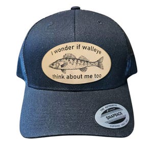 May include: A navy blue trucker hat with a mesh back and a curved brim. The hat features a tan oval patch with a detailed illustration of a walleye fish and the text "I wonder if walleye think about me too."