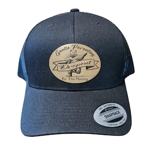 May include: A navy blue trucker hat with a curved brim and mesh back. The hat features a wooden patch with the words "Gentle Parenting Dropout" and a bear illustration. The hat has an adjustable snapback closure.