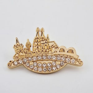 Könnte beinhalten: Eine goldfarbene Metallstifte mit einer detaillierten Silhouette einer Stadtlandschaft mit einer Brücke und einem Turm. Die Stifte ist mit klaren Strasssteinen verziert.