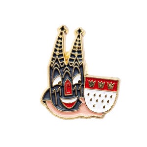 Könnte beinhalten: Goldfarbene Emaille-Anstecknadel mit einem Cartoon-Kirchengebäude mit einem lächelnden Gesicht und einem rot-weißen Schild mit drei goldenen Kronen.