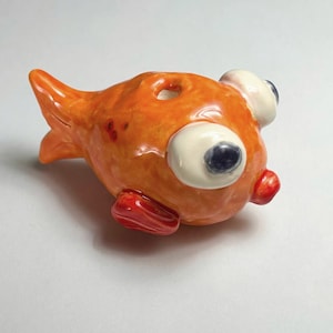 Könnte beinhalten: Orangefarbene Keramikfigur eines Fisches mit großen weißen Augen und roten Flossen. Der Fisch hat ein Loch oben auf dem Kopf.
