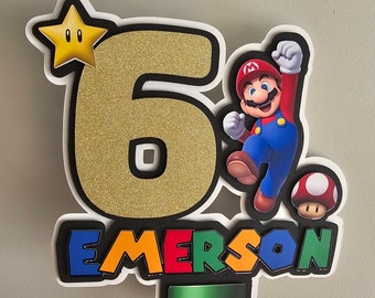 Könnte beinhalten: Ein goldener Glitzer-Kuchentopper mit der Zahl sechs, einem Mario-Charakter, einem Pilz und dem Namen "EMERSON" in bunten Buchstaben. Der Topper befindet sich auf einem weißen Hintergrund.