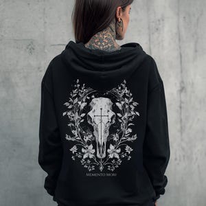 Pode incluir: Moletom preto com um design de caveira branca, cruz e elementos florais. As palavras "Memento Mori" são impressas abaixo. O moletom é escuro e tem capuz.