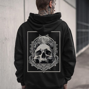 May include: Black hooded sweatshirt featuring a detailed skull graphic on the back. The skull is framed within an ornate border, with intricate floral and vine-like elements. The design is in shades of gray and white.
