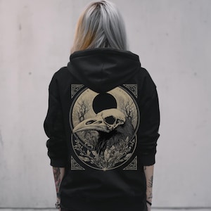 Puede incluir: Sudadera con capucha negra con una ilustración detallada de una calavera de cuervo dentro de un diseño circular. La obra de arte incluye elementos botánicos y una luna creciente, creando una estética gótica. La sudadera es oscura y tiene capucha.