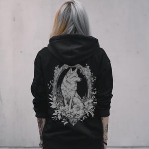 Pode incluir: Moletom preto com uma ilustração branca de um lobo dentro de uma moldura floral nas costas. O design é detalhado e central. O moletom tem capuz e um estilo clássico e simples.