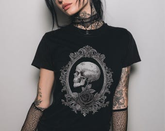 May include: Black t-shirt featuring a detailed grayscale skull design within an ornate oval frame, with a rose at the bottom. The design is centered on the shirt, creating a gothic or alternative aesthetic.
