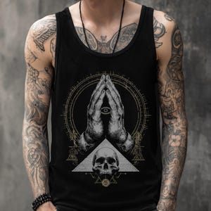 Könnte beinhalten: Ein schwarzes Tanktop mit einem Grafikdruck von zwei in Gebetshaltung zusammengelegten Händen mit einem Auge in der Mitte, platziert über einem menschlichen Schädel und geometrischen Linienelementen in Weiß-, Grau- und Goldtönen.