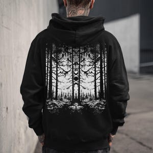 May include: Black hooded sweatshirt with a white graphic of a forest scene on the back. The design features tall trees, branches, and a moon phase symbol. The sweatshirt is made of a soft material and has a hood and front pocket.