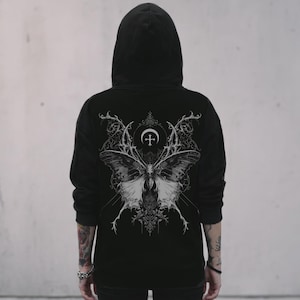 May include: A black hooded sweatshirt featuring a large, detailed white graphic on the back. The design depicts a symmetrical butterfly with intricate, ornate patterns, topped with a crescent moon and a cross symbol, set against a dark background.