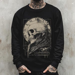 May include: Black long-sleeve shirt featuring a detailed skull graphic with a clock design and the text "TIME DEVOURS". The design is in shades of beige and gray, creating a vintage aesthetic. The shirt is a crew neck style.