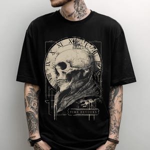 以下が含まれることがあります： 黒いTシャツ。詳細なスカルグラフィック、時計のデザイン、そして「TIME DEVOURS」の文字が特徴です。デザインはグレーとベージュの色合いです。Tシャツはクルーネックです。