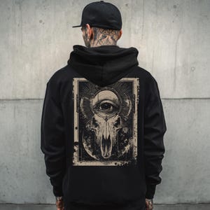 Puede incluir: Sudadera con capucha negra con un gráfico en la espalda. El diseño incluye una calavera con un ojo, rodeada de líneas radiantes, dentro de un marco rectangular. La sudadera tiene capucha y un corte holgado.