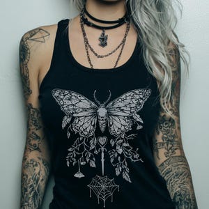 May include: Black tank top with a detailed white moth graphic. The design includes a skull, floral elements, a dagger, a heart, and a spiderweb. The tank top is sleeveless and made of a soft material.