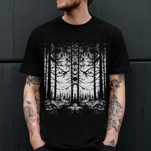 以下が含まれることがあります： 黒いTシャツ。白いグラフィックは、高い木々と中心の円形デザインが特徴の森のシーンを描いています。デザインは対称的で、鏡面効果を生み出しています。Tシャツはクルーネックです。