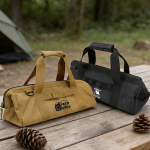 May include: Two canvas duffel bags, one tan and one black, with top handles and side pockets, sit on a wooden picnic table. The tan bag has the text "Pack Bags" printed on it. A pinecone rests on the table.