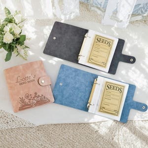 May include: Three seed books in the image. One is pink with the text "Lotte's SEED BOOK" and a floral design. The other two are open, one gray and one blue, each with a "SEEDS" label and ring binder.