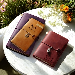 May include: Three leather journals in shades of purple, tan, and burgundy, arranged on a white surface. The tan journal has the name "Mia" and a decorative design. The burgundy journal is engraved with the initials "S|L". Each journal has a strap closure.