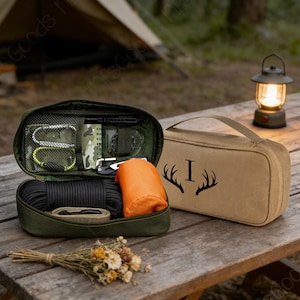 May include: A green survival kit containing a compass, carabiners, and rope, alongside a tan bag with a handle and a deer antler design. A lit lantern and a tent are visible in the background, set on a wooden table.